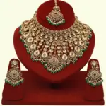 Traditional matte gold necklace set with ruby stones, green beads, intricate carvings, earrings, and maang tikka displayed on red stand, Ishwar Arts and Crafts