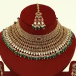Premium matte gold bridal necklace set with ruby and green stones, heavy choker design, hanging green beads, matching earrings and maang tikka displayed on red stand