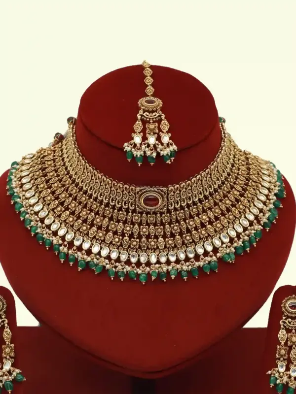 Premium matte gold bridal necklace set with ruby and green stones, heavy choker design, hanging green beads, matching earrings and maang tikka displayed on red stand
