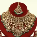 Traditional matte gold necklace set with ruby stones, green beads, intricate carvings, earrings, and maang tikka displayed on red stand