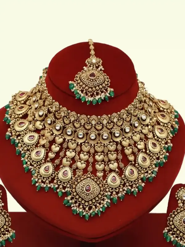 Traditional matte gold necklace set with ruby stones, green beads, intricate carvings, earrings, and maang tikka displayed on red stand
