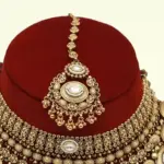 Heavy matte gold brass bridal necklace set with kundan stones, earrings, and maang tikka displayed on red jewelry stand