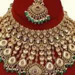 Traditional matte gold necklace set with ruby stones, green beads, intricate carvings, earrings, and maang tikka displayed on red stand, Ishwar Arts and Crafts