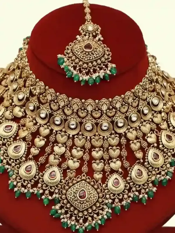 Traditional matte gold necklace set with ruby stones, green beads, intricate carvings, earrings, and maang tikka displayed on red stand, Ishwar Arts and Crafts
