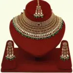 Premium matte gold bridal necklace set with ruby and green stones, heavy choker design, hanging green beads, matching earrings and maang tikka displayed on red stand