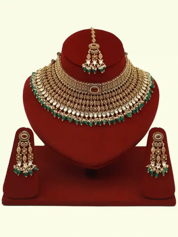 Premium matte gold bridal necklace set with ruby and green stones, heavy choker design, hanging green beads, matching earrings and maang tikka displayed on red stand