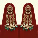 Premium matte gold bridal necklace set with ruby and green stones, heavy choker design, hanging green beads, matching earrings and maang tikka displayed on red stand