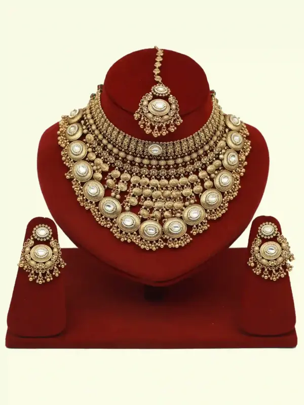 Heavy matte gold brass bridal necklace set with kundan stones, earrings, and maang tikka displayed on red jewelry stand