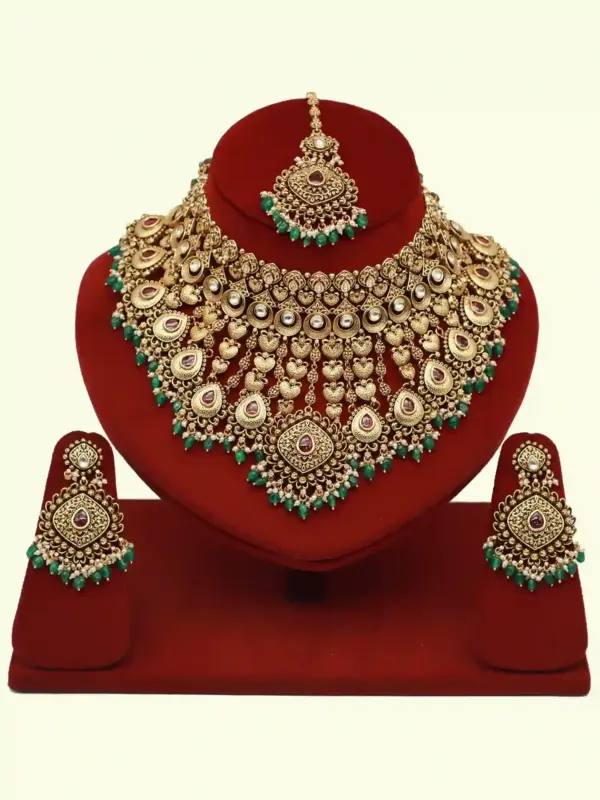 Traditional matte gold necklace set with ruby stones, green beads, intricate carvings, earrings, and maang tikka displayed on red stand